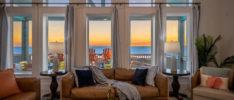 A spacious living room with a large window overlooking a balcony. The room features a brown leather sofa, a coffee table with a bottle of wine, and a view of the sunset.