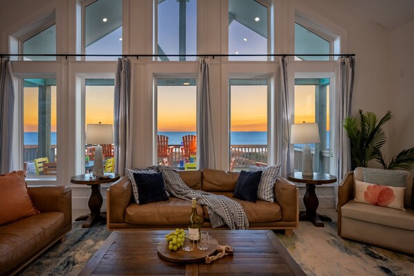 A spacious living room with a large window overlooking a balcony. The room features a brown leather sofa, a coffee table with a bottle of wine, and a view of the sunset.