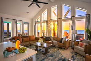 A spacious living room with a large window overlooking the ocean, a ceiling fan, and a dining area with a table set for two.