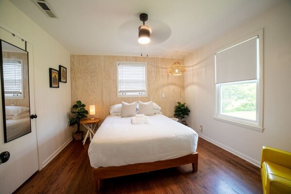 A bedroom with a wooden bed, two bedside tables with lamps, a large window with blinds, and a ceiling fan.