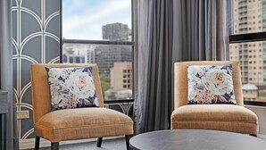 Deux fauteuils ornés de coussins à motifs floraux, une table ronde et une vue sur le paysage urbain, visible à travers de grandes fenêtres.