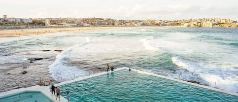 Zona costera para nadar con piscina contigua al mar, una playa al fondo y un paisaje urbano al otro lado del agua.