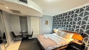 Habitación de hotel con cama, mesita de noche, lámpara y un escritorio pequeño con silla.