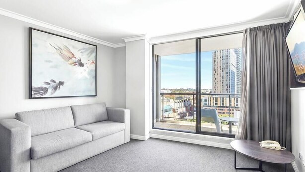 A modern hotel room with a grey sofa, a large window with a view of buildings, and a framed artwork on the wall. - KozyGuru | Central Sydney | 2 Bed 2 Bath City View (Haymarket)