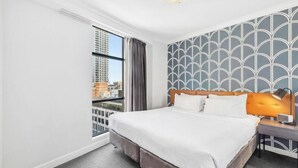 A hotel room with a large bed, a nightstand, a lamp, a window with curtains, and a patterned wall. - KozyGuru | Central Sydney | 2 Bed 2 Bath City View (Haymarket)