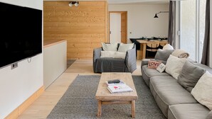 Ein modernes Wohnzimmer mit Flachbildfernseher, Holztisch, grauem Sofa und Essbereich im Hintergrund.