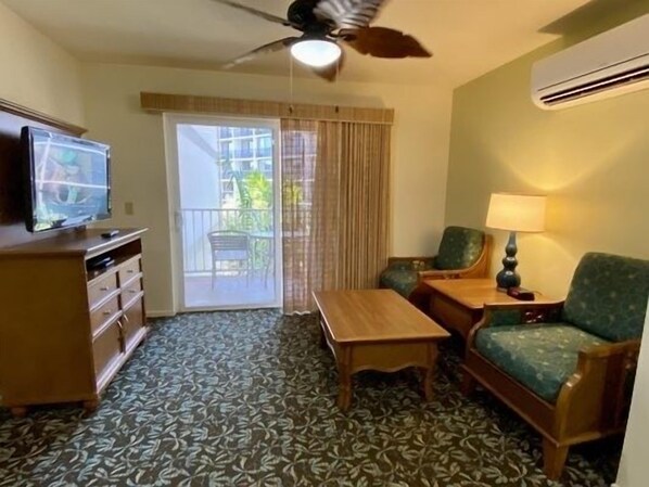A hotel room with a ceiling fan, a television on a wooden cabinet, a small table, a green armchair, a lamp, and a patterned carpet.