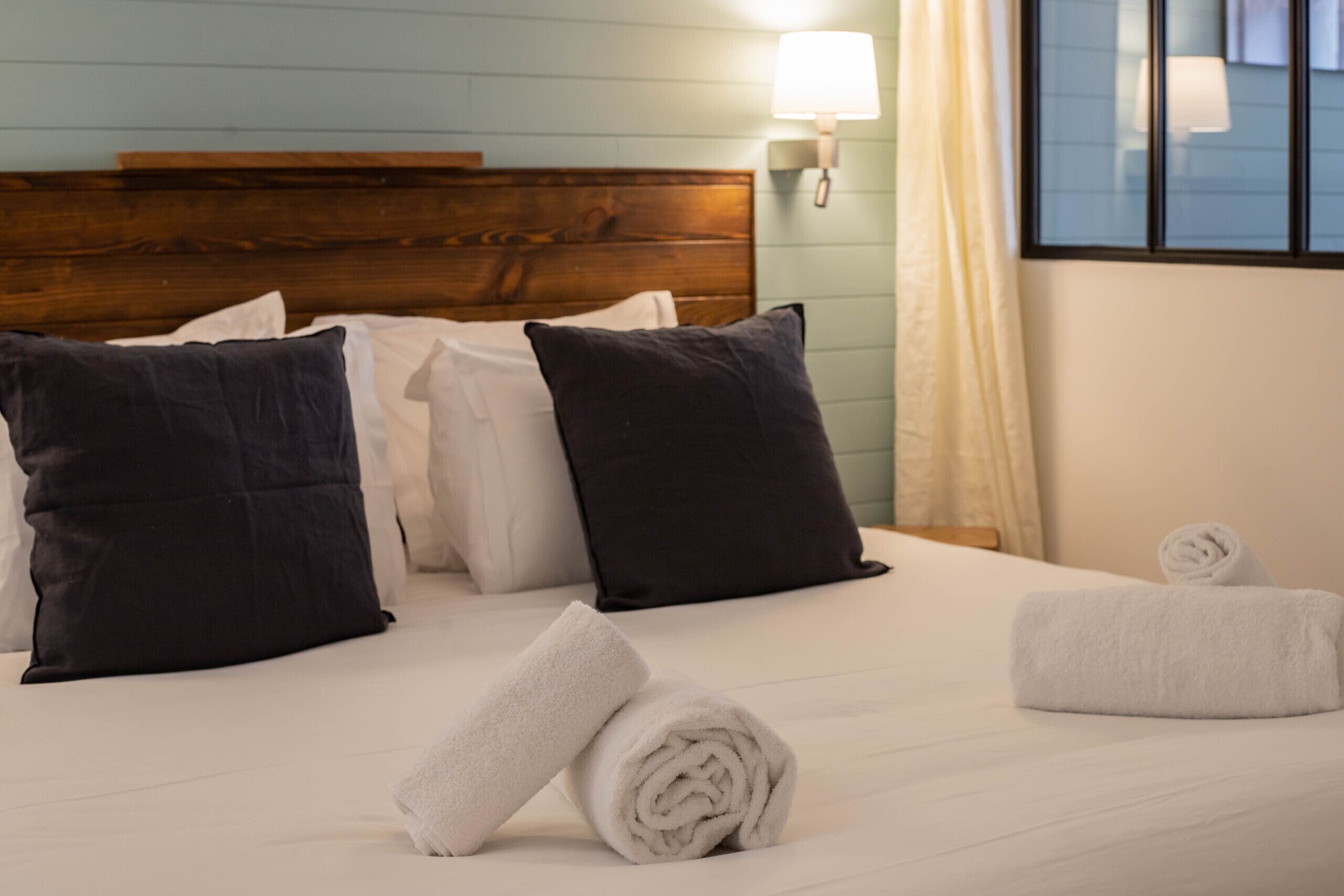 Een netjes opgemaakt bed met witte en zwarte kussens, een opgerolde witte handdoek en een raam met uitzicht.