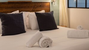 A neatly made bed with white and black pillows, a rolled white towel, and a window with a view of the outside.