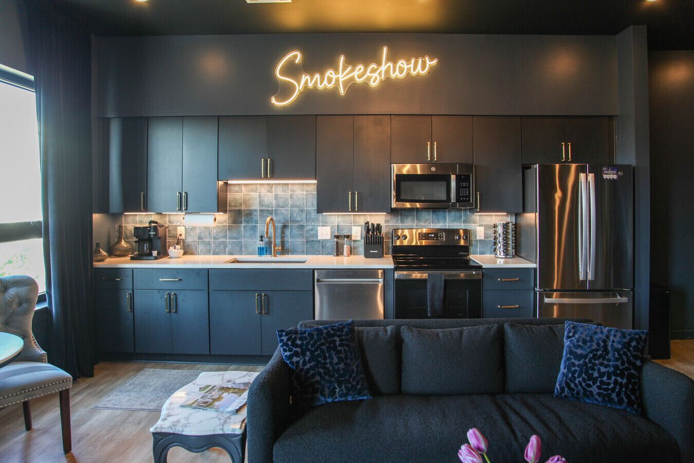 A modern kitchen with dark cabinetry, a neon sign reading "Smoke show", stainless steel appliances, and a cozy seating area with a dark sofa and patterned cushions.