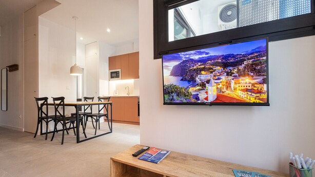 Un salon moderne avec un téléviseur à écran plat fixé au mur, une table basse en bois sur laquelle sont posés des livres et une télécommande, et un coin repas avec une table et des chaises.