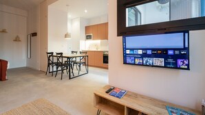 Un salon moderne avec un téléviseur à écran plat fixé au mur, une table à manger en bois avec des chaises, et une cuisine en arrière-plan.