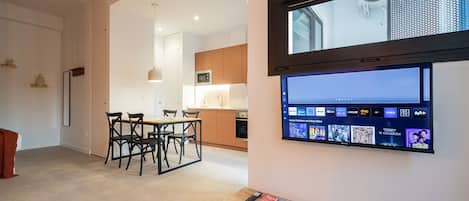 A modern living room with a flat-screen TV mounted on the wall, a wooden dining table with chairs, and a kitchen in the background.