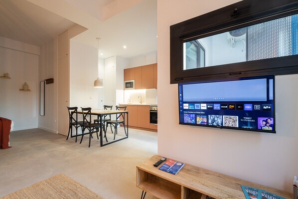 A modern living room with a flat-screen TV mounted on the wall, a wooden dining table with chairs, and a kitchen in the background.