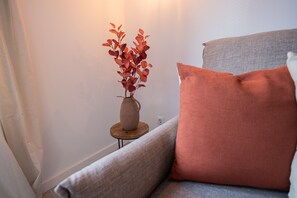 A corner of a room with a grey sofa, a wooden side table, and a vase with red leaves.