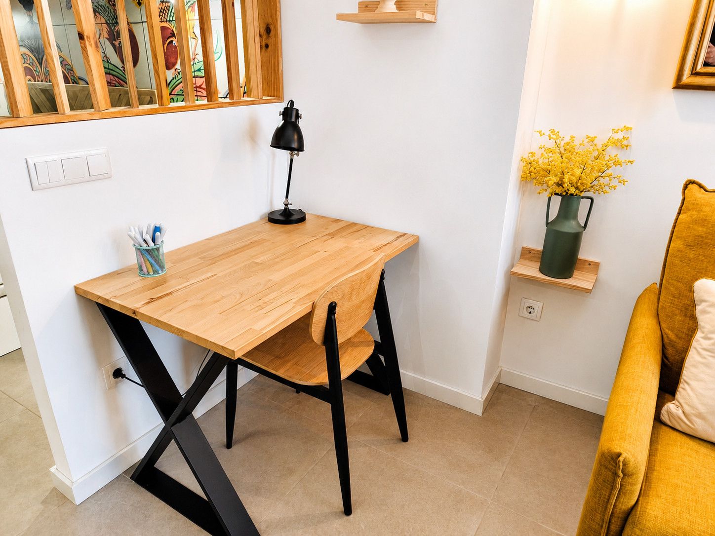 Een knus kamertje met een houten bureau, een zwarte stoel, een klein plankje met een vaas en een gele bank.