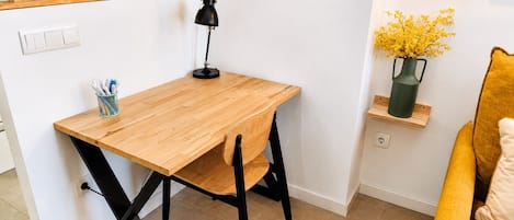 Een knus kamertje met een houten bureau, een zwarte stoel, een klein plankje met een vaas en een gele bank.