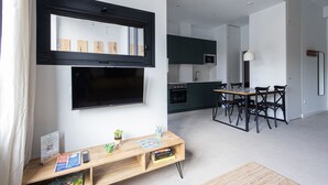 A modern living room with a flat-screen TV, wooden coffee table, and books. A kitchen with a microwave and stove is visible in the background.