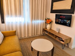 Un salon comprenant un canapé, une table basse, un téléviseur fixé au mur diffusant Netflix, et un vase avec des fleurs placé sur une table d’appoint.
