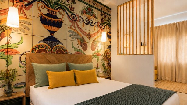 A hotel room with a bed, a decorative wall, a wooden headboard, a bedside table, and a window with curtains.