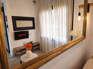 Ein Zimmer mit Fernseher, der 'NETFLIX' anzeigt, einer weißen Vase mit Blumen, einer Lampe und einem Spiegel, der das Zimmer widerspiegelt.