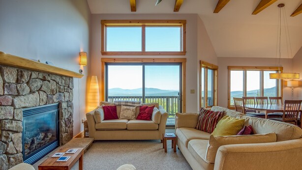 A cozy living room with a stone fireplace, large windows, and a dining area.