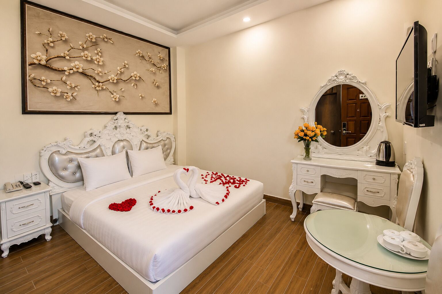 A hotel room with a large bed, a vanity with a mirror, a small table, and a wall-mounted TV.