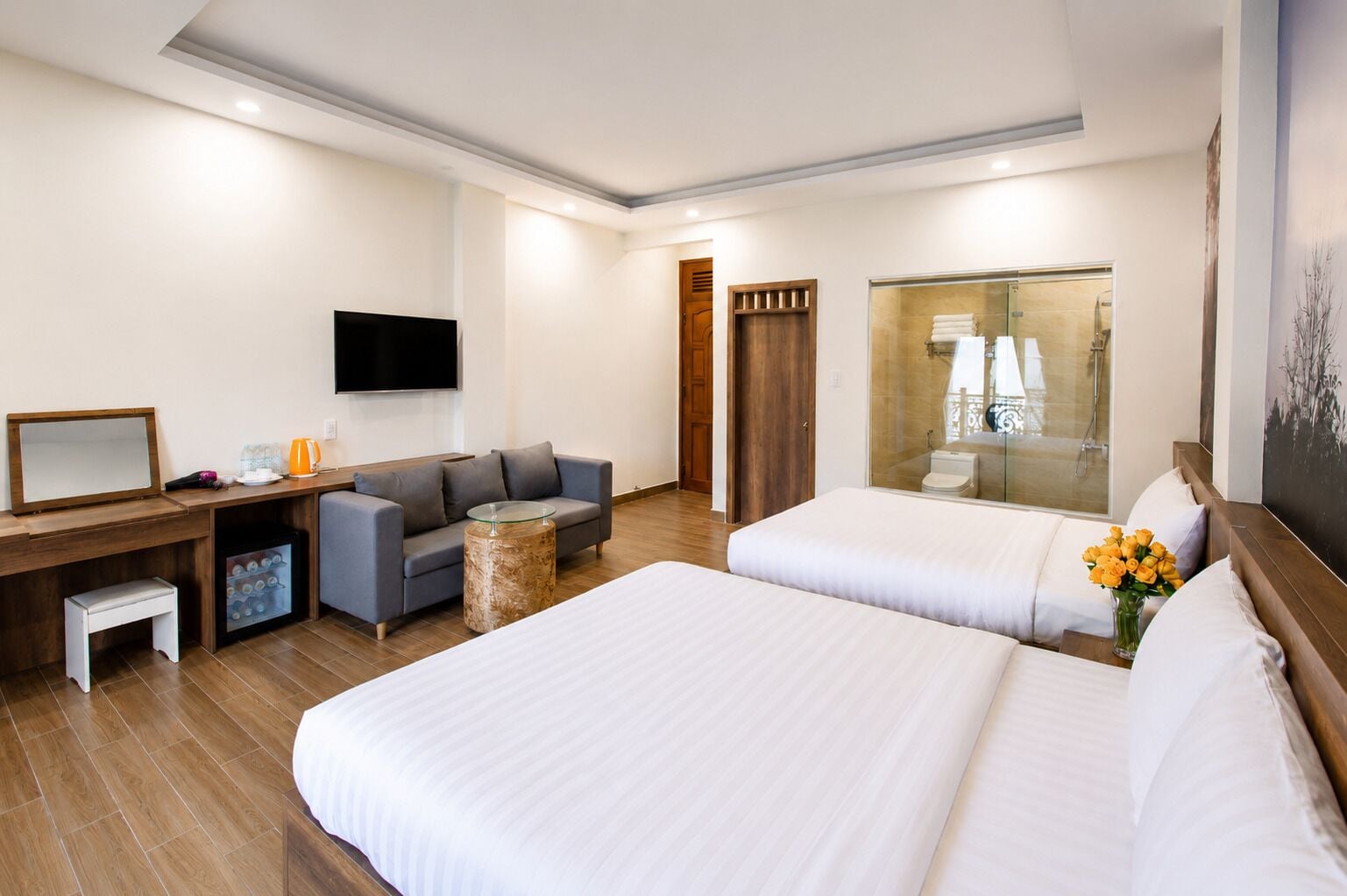 A hotel room with two beds, a sofa, a desk, a TV, and a bathroom visible through a glass door.