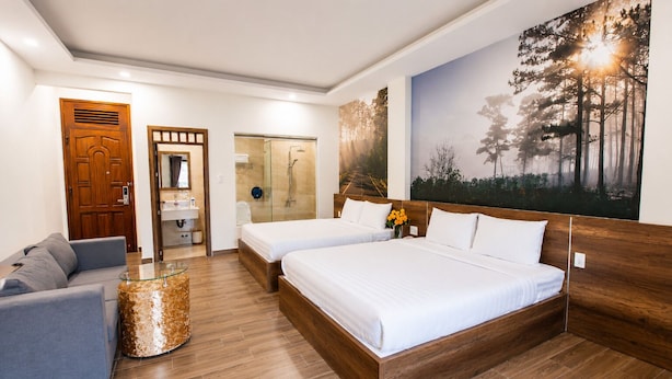 A hotel room with a wooden floor, two beds, a gray sofa, a glass coffee table, a bathroom with a glass shower, and a forest-themed wall mural. - Lien's Hotel Da Lat (Xuan Huong - Da Lat)