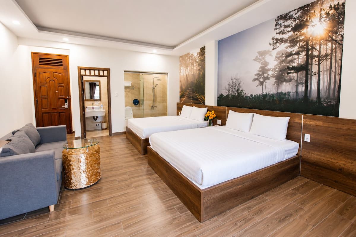 A hotel room with a wooden floor, two beds, a gray sofa, a glass coffee table, a bathroom with a glass shower, and a forest-themed wall mural.
