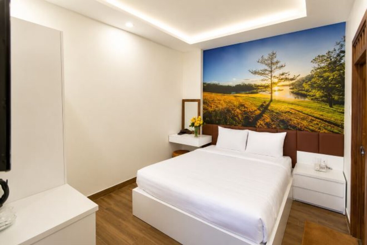 Quarto com cama, mesa de cabeceira, cômoda, espelho e um painel com pintura de paisagem.