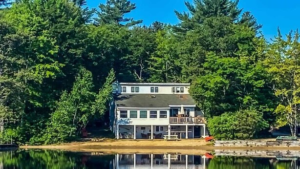 A two-story waterfront house with a dock, surrounded by dense trees. - Lake Home with Private Beach and Hot Tub (Ossipee)