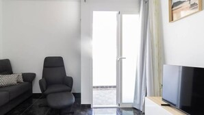 Un salon moderne avec un canapé, un fauteuil noir, un téléviseur à écran plat posé sur un meuble en bois et une porte-fenêtre coulissante donnant sur un espace extérieur.