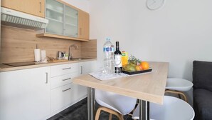 Une cuisine de taille réduite, dotée d’un plan de travail en bois, d’un évier et de placards. Il y a une horloge au mur et une table sur laquelle sont disposés des fruits et des bouteilles.