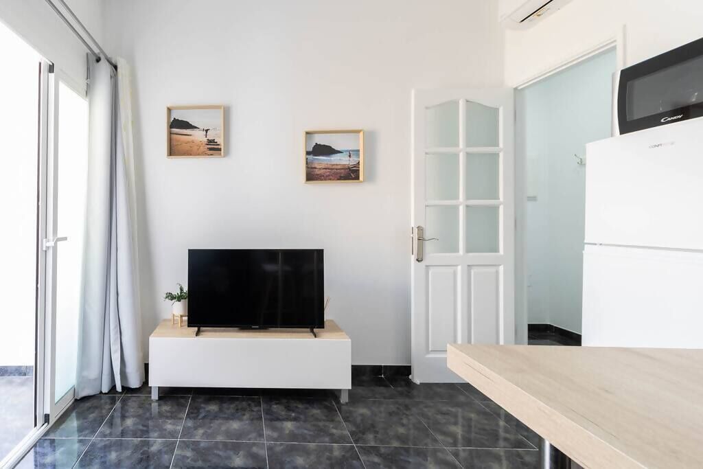 A modern living room with a flat-screen TV on a white console, two framed pictures on the wall, and a sliding glass door leading to a balcony.
