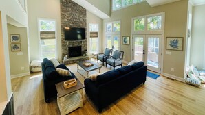 A spacious living room with a stone fireplace, large windows, and a ceiling fan.