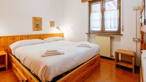 A bedroom with a wooden bed, a window with curtains, a radiator, and a small wooden stool. - Apartment Apt 18 by Interhome (Canazei)