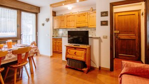 A cozy living room with a wooden TV stand, a dining table with chairs, and a kitchen area with wooden cabinets and a tiled backsplash. - Apartment Apt 18 by Interhome (Canazei)