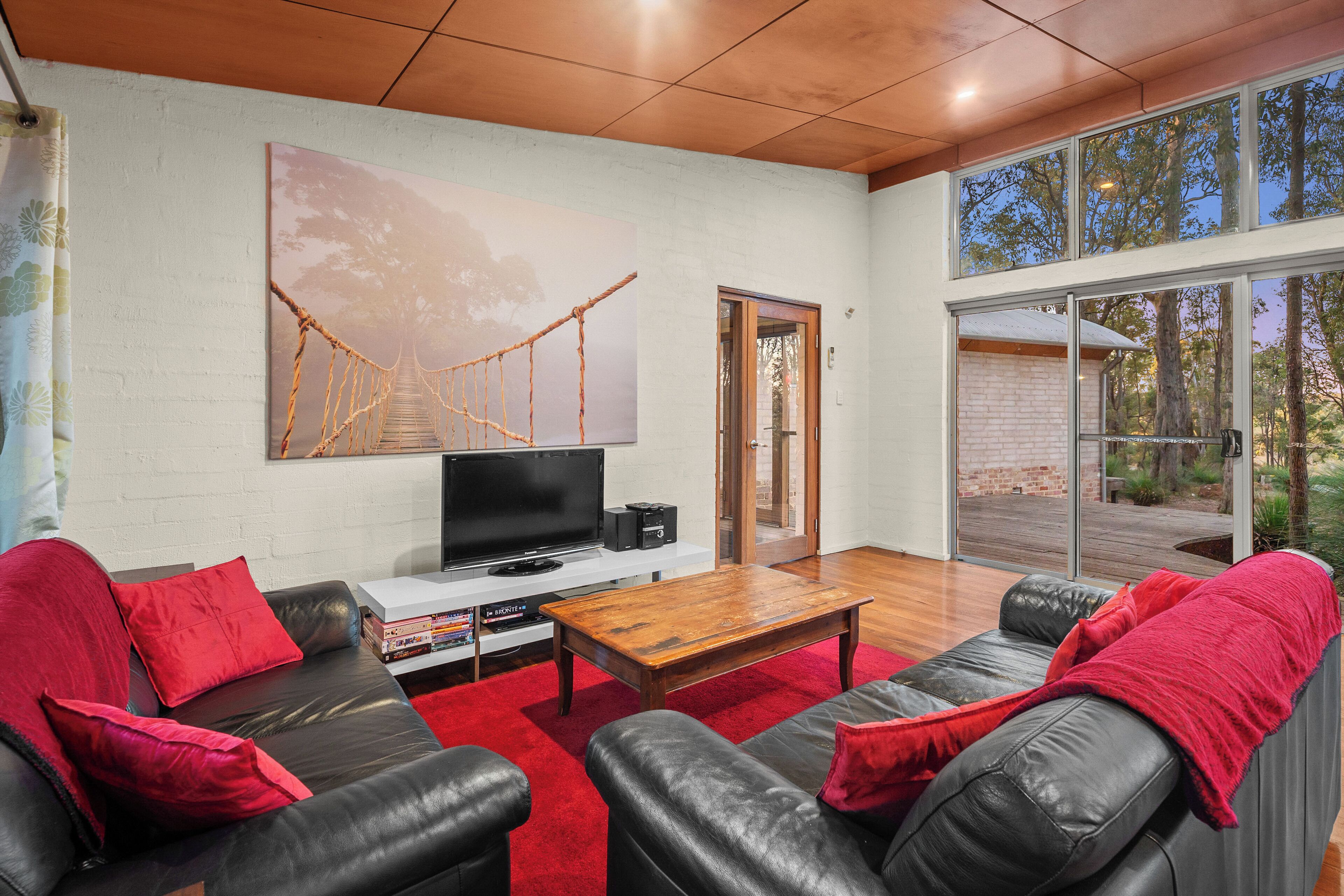A modern living room with a large window, a wooden coffee table, black leather sofas, and a red rug.