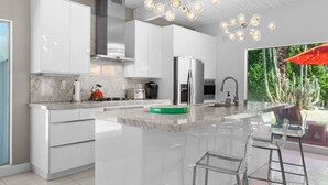 A modern kitchen with white cabinetry, a central island, and a large window overlooking a garden. - Lush, secluded desert oasis w/pool & EV charger (Palm Springs)