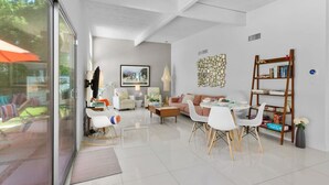 A modern living room with a glass door leading to a patio, a dining area with white chairs, and a bookshelf. - Lush, secluded desert oasis w/pool & EV charger (Palm Springs)