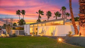 A modern house with a large glass facade, surrounded by palm trees and a well-maintained lawn. - Lush, secluded desert oasis w/pool & EV charger (Palm Springs)
