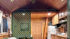 A cozy room with a wooden ceiling, a green patterned partition, a sofa with cushions, a small wooden table, and a kitchen area with wooden cabinets and a refrigerator. - Private Romantic Woodland Getaway- sauna, hot tub, outdoor culinary bar, gardens (Vader)