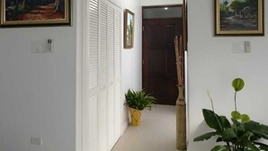 Un couloir aux murs blancs, orné de deux tableaux encadrés, avec une plante en pot et une porte au fond.