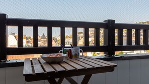 Une table de pique-nique en bois, sur laquelle sont disposés un saladier rempli de viennoiseries et une tasse, le tout placé devant la rambarde d’un balcon donnant sur un paysage urbain.