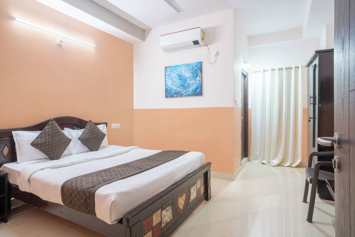 Una cama bien hecha con cabecera, una mesita de noche, aire acondicionado de pared, un cuadro y una puerta con cortinas.