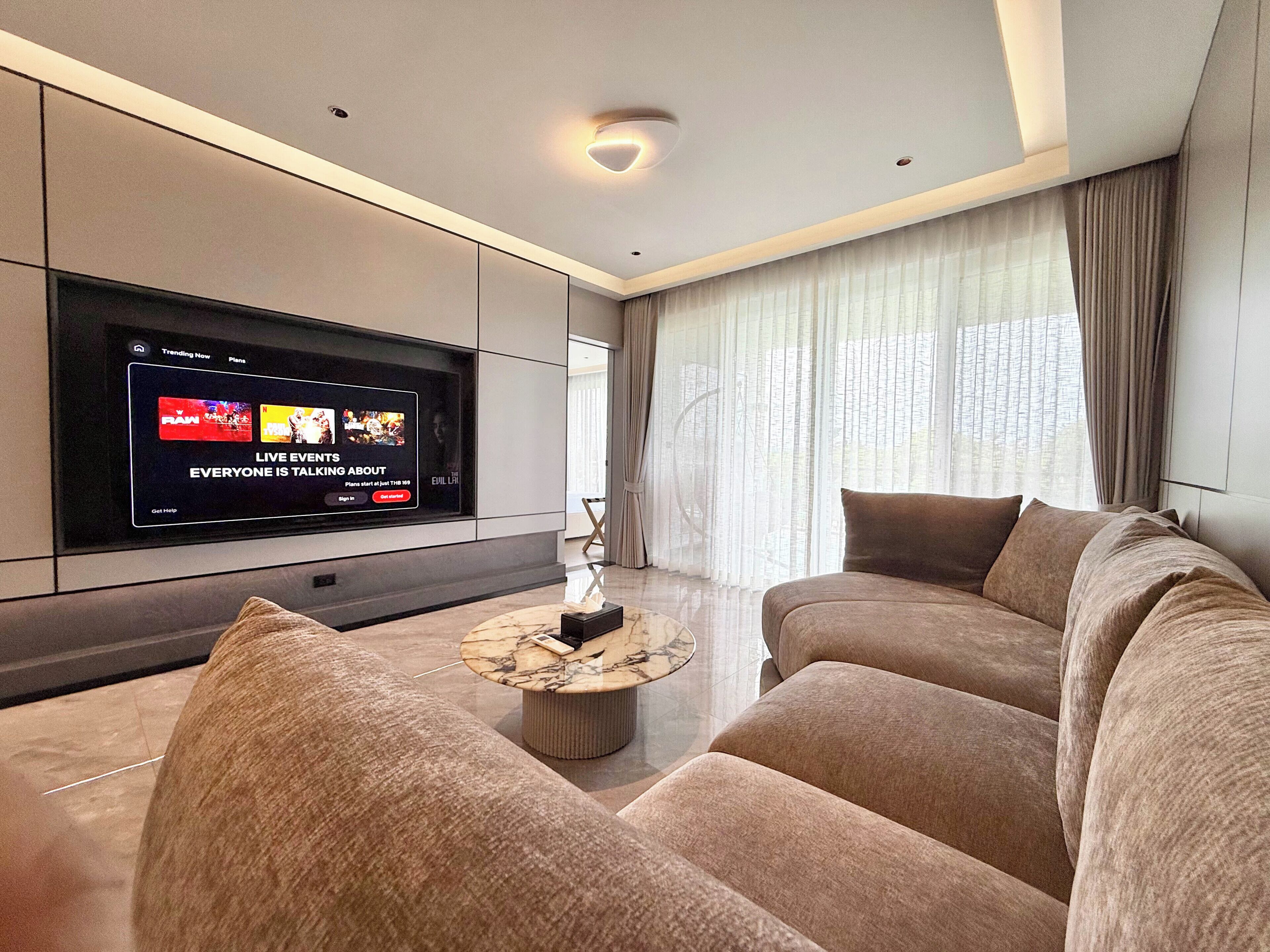 A modern living room with a large flat-screen TV, a marble coffee table, and plush sofas.