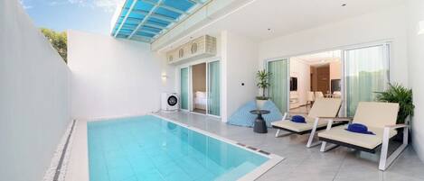 A modern indoor pool area with a glass ceiling, a lounge with chairs, and a view into a living area.