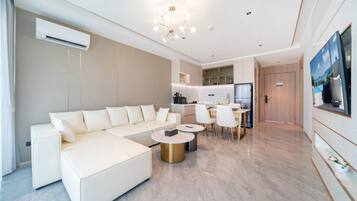 A modern living room with a white sectional sofa, a dining area with a table and chairs, a kitchen with a refrigerator, and a large painting on the wall.