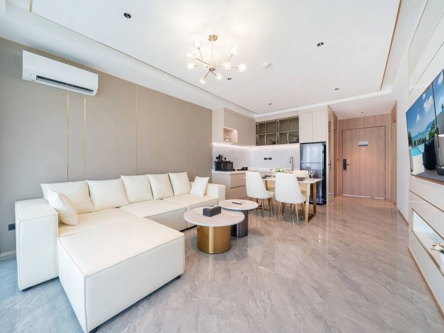 A modern living room with a white sectional sofa, a dining area with a table and chairs, a kitchen with a refrigerator, and a large painting on the wall.
